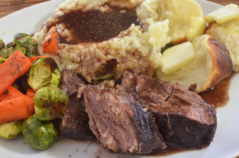 A beef chuck roast braising in a Dutch oven with carrots, onion, and herbs in a partially reduced liquid.
